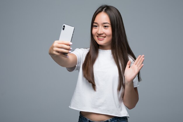 Mujer asiática joven sonriente que toma un selfie con el teléfono móvil sobre fondo gris aislado de la pared