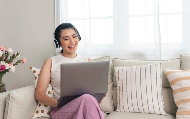 Mujer asiática joven sonriente que usa una computadora portátil mientras se relaja en un sofá en casa trabajando desde casa