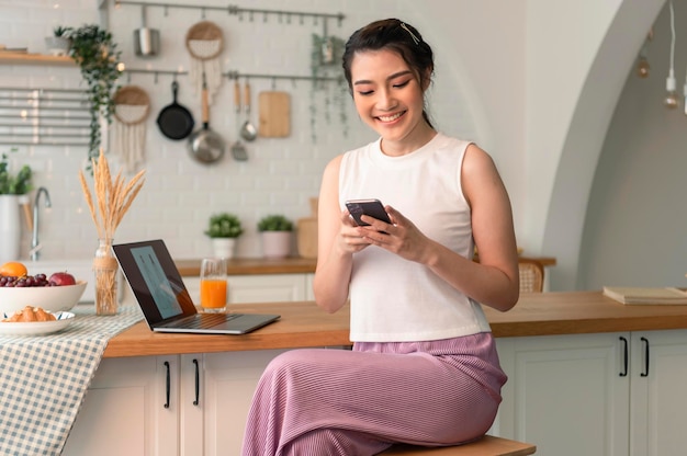 Mujer asiática joven sonriente que usa el teléfono móvil mientras está sentada en la cocina en casa con una computadora portátil