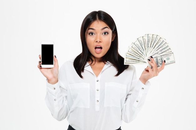 Foto gratuita mujer asiática joven sorprendida que sostiene el dinero que muestra la exhibición del teléfono.