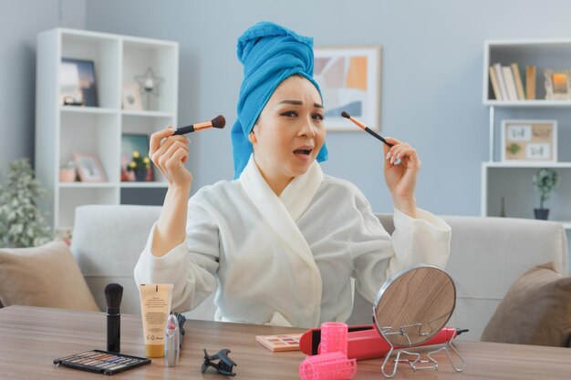 Mujer asiática joven con una toalla en la cabeza sentada en el tocador en el interior de la casa aplicando sombras de ojos o colorete con espejo haciendo rutina de maquillaje matutino con aspecto confundido