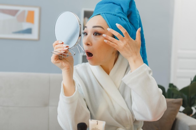 Mujer asiática joven con una toalla en la cabeza sentada en el tocador en el interior de la casa mirándose en el espejo confundida tocando su ojo haciendo la rutina de maquillaje matutino