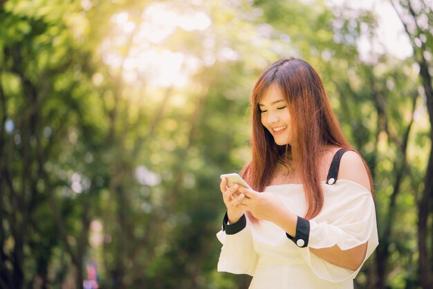 La mujer asiática linda está leyendo el mensaje de texto agradable en el teléfono móvil mientras que se sienta en parque en día de primavera caliente, hembra magnífica que escucha la música en auriculares y que busca la información en el teléfono celular.