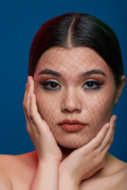 Mujer asiática con maquillaje completo, delgada red que cubre la cara y las manos en las mejillas posando en el estudio