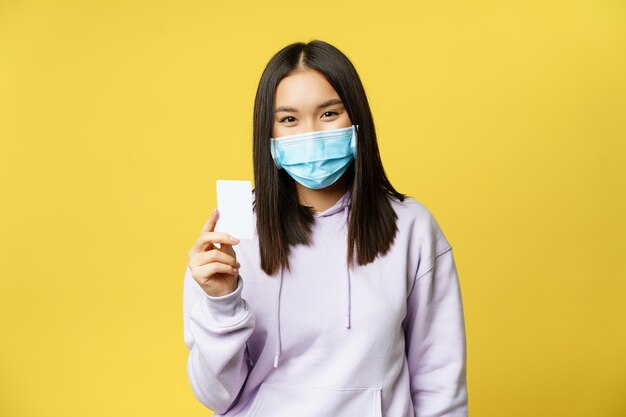 Mujer asiática en mascarilla médica mostrando pase de tarjeta de crédito de pie sobre fondo amarillo