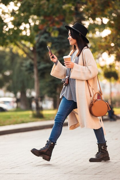 Mujer asiática moderna con smartphone y sosteniendo café para llevar mientras camina en el parque al aire libre