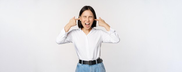 Una mujer asiática molesta que siente incomodidad por un ruido fuerte y molesto cierra los oídos y los ojos de pie sobre fondo blanco