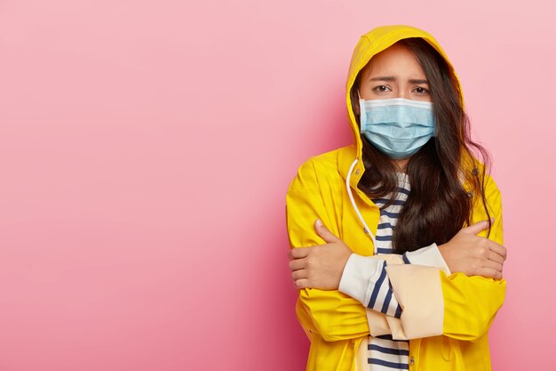 Mujer asiática molesta tiembla de frío, tiene el virus transmitido a través de gotitas de aire, usa máscara médica protectora, impermeable amarillo con capucha