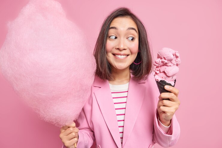 Foto gratuito mujer asiática morena de aspecto agradable mira apetitoso helado de cono sostiene hilo de caramelo en un palo disfruta de postres de verano come comida chatarra vestida con chaqueta rosa posa interior tiene caminar en el parque