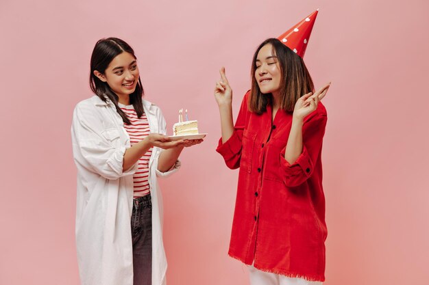 Mujer asiática morena en camisa larga presenta pastel de cumpleaños a su amiga Niña en sombrero de fiesta hace deseo cruza los dedos sobre fondo rosa