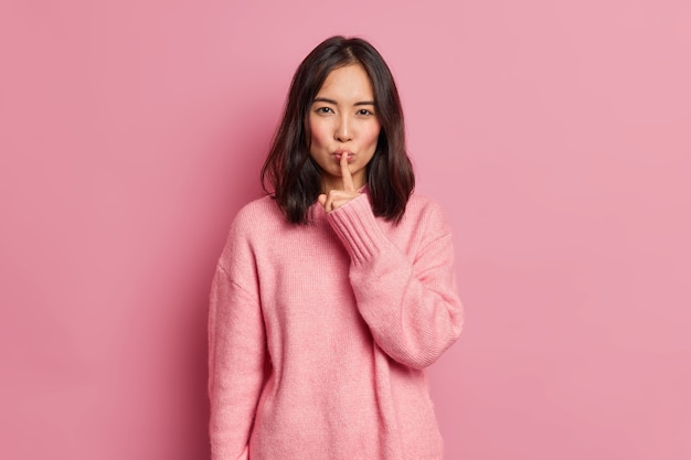 La mujer asiática morena misteriosa seria presiona el dedo índice a los labios hace un gesto de silencio dice que el secreto pide estar callado usa poses de jersey de manga larga