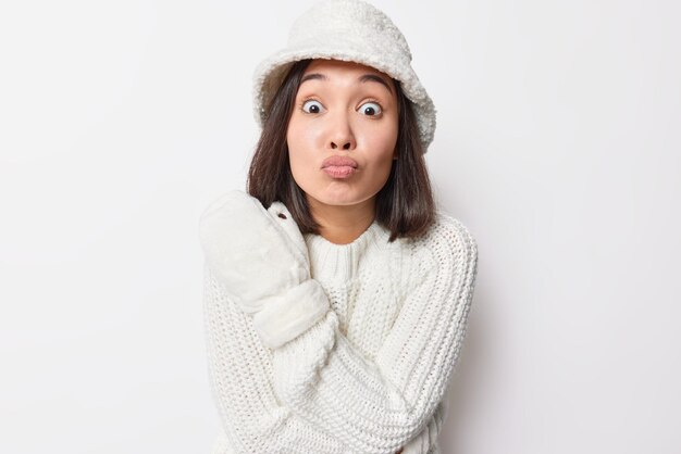 La mujer asiática morena tierna y romántica mantiene los labios doblados y quiere besarte, usa suéter de punto con sombrero de piel blanca y mitones disfruta del invierno aislado sobre fondo blanco. Expresiones del rostro humano