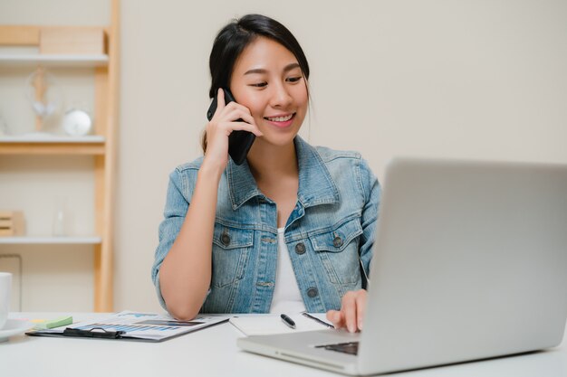 Mujer asiática del negocio elegante hermoso en la ropa de sport elegante que trabaja en el ordenador portátil y que habla en el teléfono mientras que se sienta en la tabla en oficina creativa.
