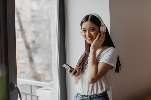 La mujer asiática de ojos marrones en la parte superior blanca mira al frente con una sonrisa, sostiene el teléfono inteligente y se pone los auriculares
