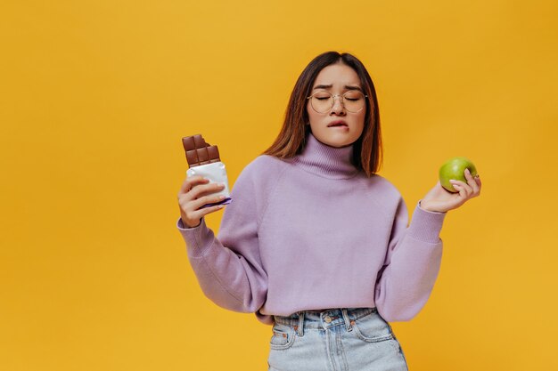 Mujer asiática de pelo corto con anteojos, suéter morado y falda de mezclilla observa su labio y trata de decidir qué elegir: manzana verde fresca o sabrosa barra de chocolate dulce