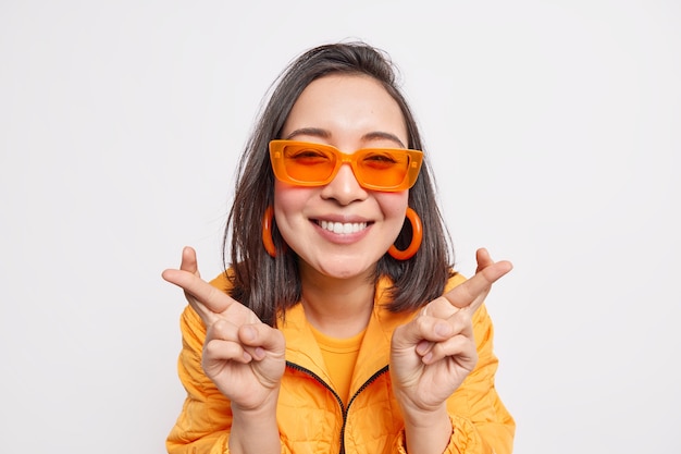 Foto gratuita la mujer asiática de pelo oscuro de moda alegre hace que el deseo mantenga los dedos cruzados espera a que el sueño se haga realidad sonríe felizmente usa aretes de gafas de sol naranjas de moda y una chaqueta aislada sobre una pared blanca