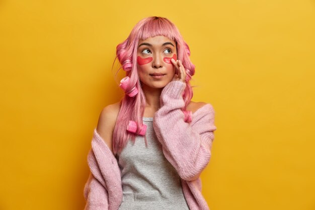 Mujer asiática de pelo rosa pensativa seria con rulos en la cabeza, toca parches para una piel suave y saludable