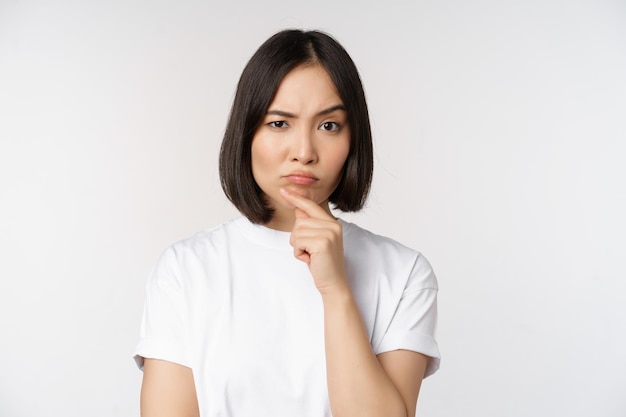 Mujer asiática pensativa mirando a un lado reflexionando sobre hacer suposiciones pensando o eligiendo algo de pie sobre fondo blanco