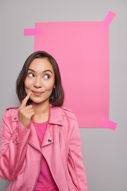 Foto gratuita mujer asiática pensativa piensa en hacer publicidad genial de productos mira el espacio de copia en blanco vestida con chaqueta casual mantiene índice cerca de labios ofrece venta o descuento