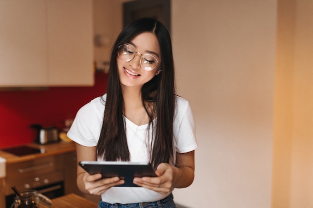 Mujer asiática positiva en camiseta blanca y gafas de moda posando con tableta de computadora