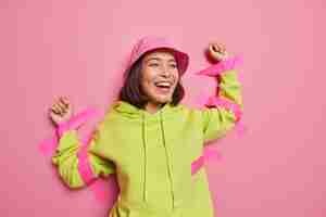 Foto gratuita la mujer asiática positiva se ríe felizmente pegada a la pared con cintas adhesivas viste panamá y la sudadera con capucha no se siente libre aislada sobre la pared rosa