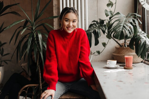 Mujer asiática positiva en suéter rojo y jeans grises se sienta a la mesa en el café