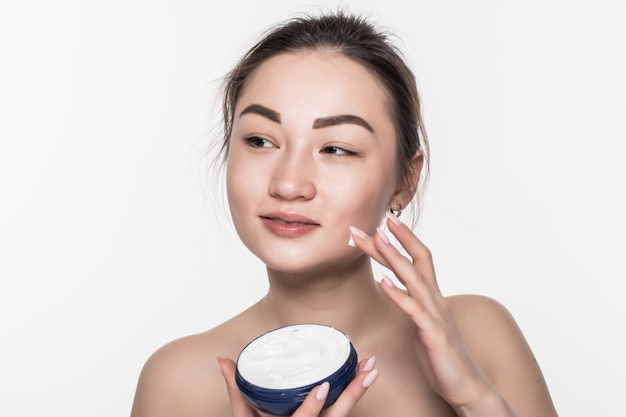Mujer asiática que aplica la crema cosmética en cara facial del cuidado de la piel en aislado en la pared blanca