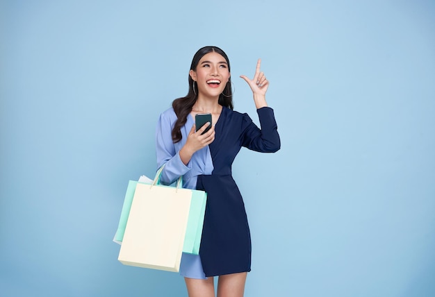 Mujer asiática que lleva bolsas de compras y compras en línea con el teléfono móvil apuntando con el dedo hacia arriba