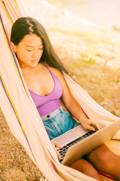 Mujer asiática que miente en hamaca y que trabaja en la computadora portátil