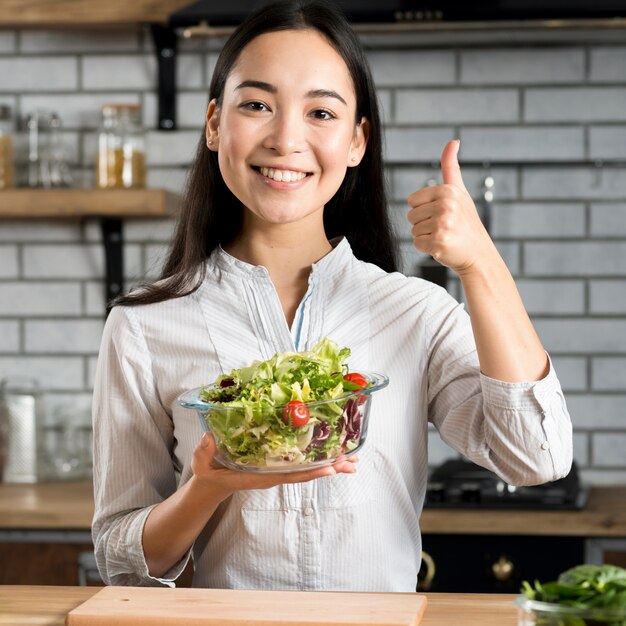 Mujer asiática que muestra el pulgar encima de la muestra con sostener la ensalada sana de las verduras en cocina