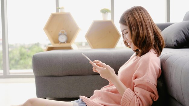 Mujer asiática que usa la tableta mientras que miente en el sofá casero en su sala de estar.