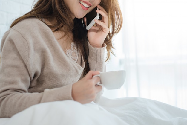 Mujer asiática que usa el teléfono inteligente en su cama mientras sostiene una taza de café por la mañana