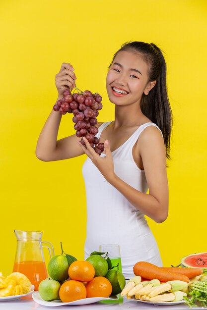 Una mujer asiática con un racimo de uvas, y sobre la mesa hay muchas frutas.