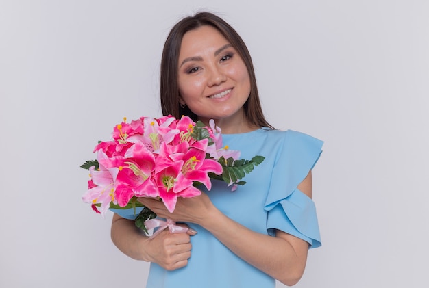 Mujer asiática con ramo de flores mirando feliz y alegre celebrando el día internacional de la mujer de pie sobre una pared blanca