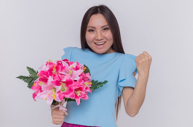 Mujer asiática con ramo de flores mirando feliz y alegre puño apretado celebrando el día internacional de la mujer de pie sobre una pared blanca