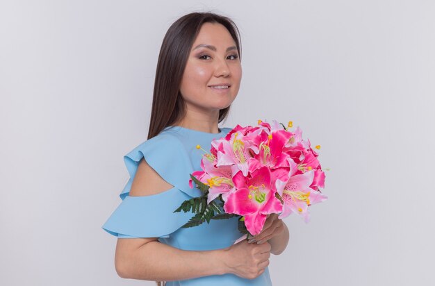 Mujer asiática con ramo de flores mirando feliz y alegre sonriendo celebrando el día internacional de la mujer de pie sobre una pared blanca