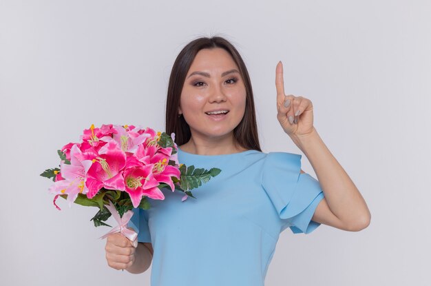 Mujer asiática con ramo de flores mirando feliz y alegre sonriendo mostrando el dedo índice celebrando el día internacional de la mujer de pie sobre una pared blanca