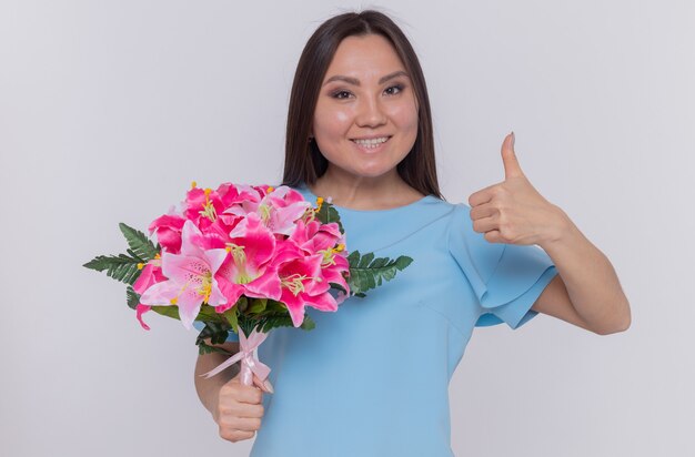 Mujer asiática con ramo de flores mirando feliz y alegre sonriendo mostrando el pulgar hacia arriba celebrando el día internacional de la mujer de pie sobre una pared blanca