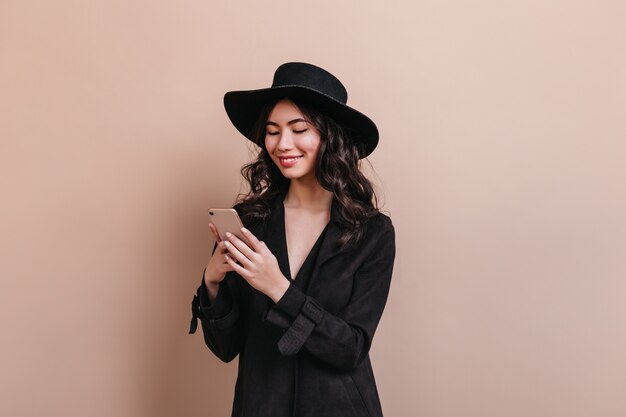 Mujer asiática rizada con smartphone. Riendo a mujer coreana en abrigo posando con gadget.