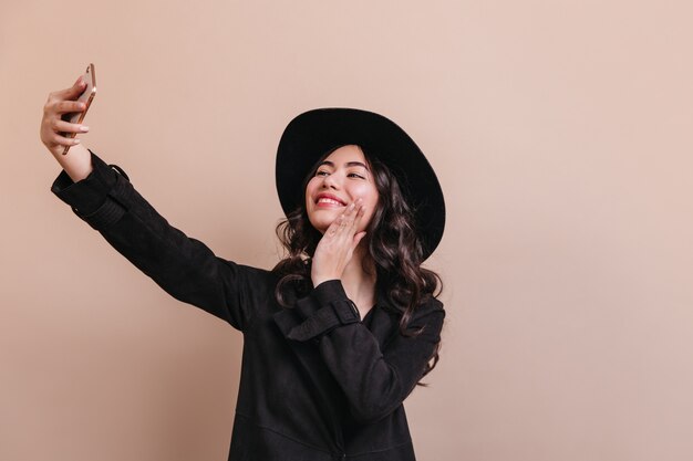 Mujer asiática romántica en abrigo tomando selfie. Foto de estudio de inspirada mujer china con sombrero.