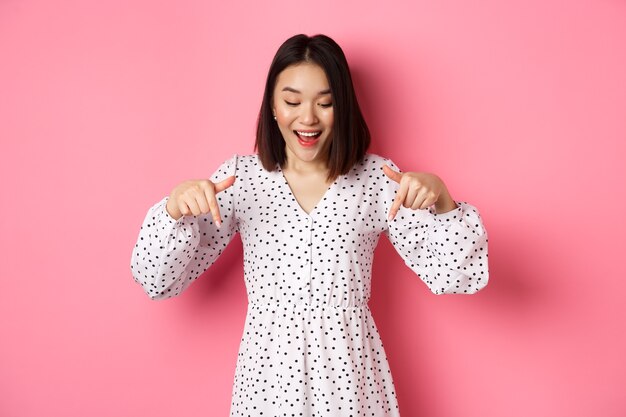 Mujer asiática romántica mirando hacia abajo, señalando con el dedo en la parte inferior, mirando con curiosidad el descuento del producto con una sonrisa feliz, de pie sobre fondo rosa