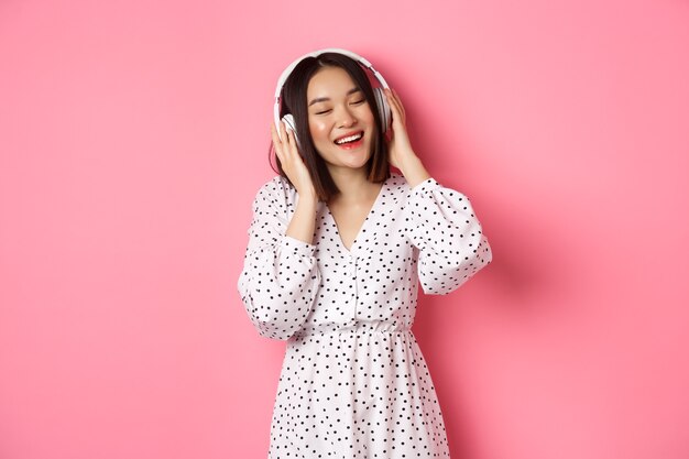 Mujer asiática romántica sonriendo feliz, escuchando música en auriculares y bailando, de pie en un vestido de moda sobre fondo rosa