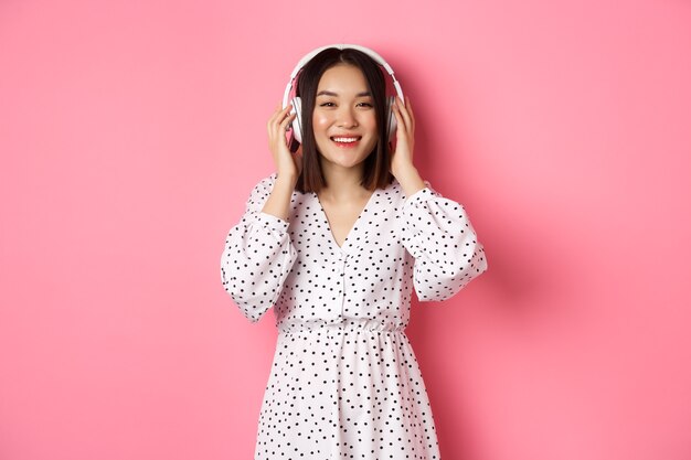 Mujer asiática romántica sonriendo feliz, escuchando música en auriculares y mirando a cámara, de pie sobre fondo rosa