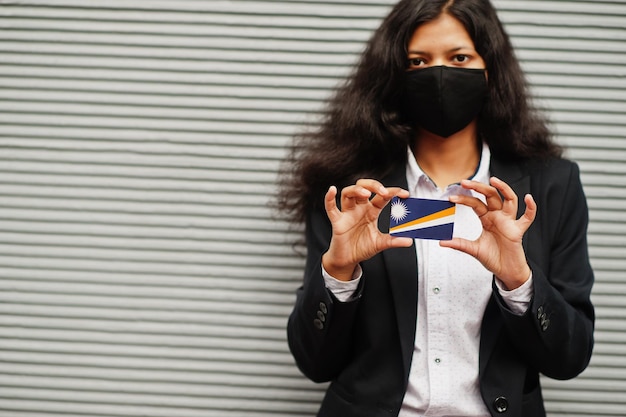 Foto gratuita mujer asiática con ropa formal y máscara protectora negra sostiene la bandera de las islas marshall a mano contra el fondo gris coronavirus en el concepto de país