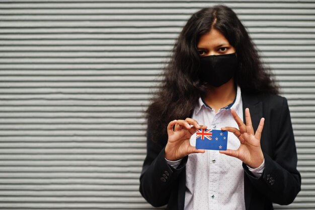 Mujer asiática con ropa formal y máscara protectora negra sostiene la bandera de Nueva Zelanda a mano contra el fondo gris Coronavirus en el concepto de país