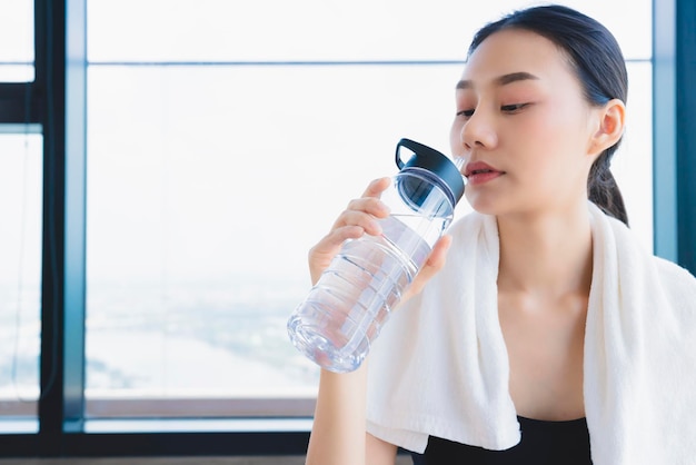 Mujer asiática saludable que se relaja en el gimnasio después de hacer ejercicio con la mano, sostenga una bebida de agua pura y una toalla blanca