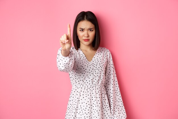 Mujer asiática seria y enojada que dice que se detenga, frunciendo el ceño y mostrando el dedo en desaprobación, prohíba algo, de pie en el vestido contra el fondo rosa.