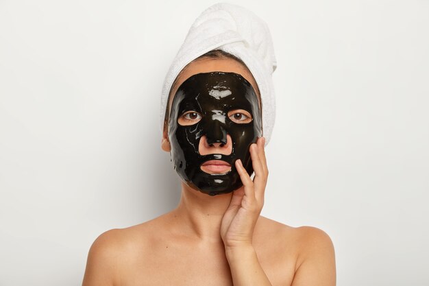 Una mujer asiática seria se somete a procedimientos cosméticos en casa, se aplica una mascarilla facial purificadora negra, se ve recta, toca suavemente la mejilla, usa una toalla blanca suave en la cabeza