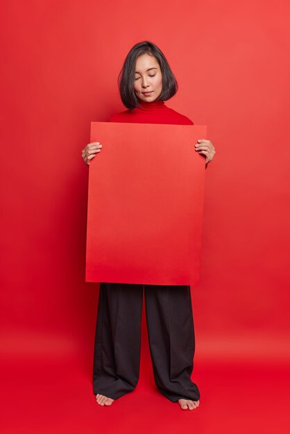 La mujer asiática seria sostiene el tablero de publicidad rojo recomienda colocar su información aquí exhibe el atuendo elegante de weras de la bandera posa contra la pared brillante del estudio