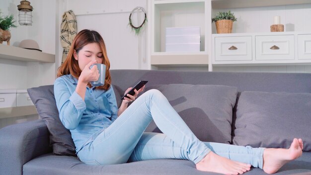 Mujer asiática sonriente atractiva hermosa que usa el smartphone que sostiene una taza de café caliente